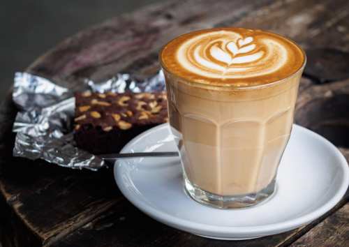 A glass of latte with a leaf design on top, served on a white plate next to a chocolate brownie on foil.