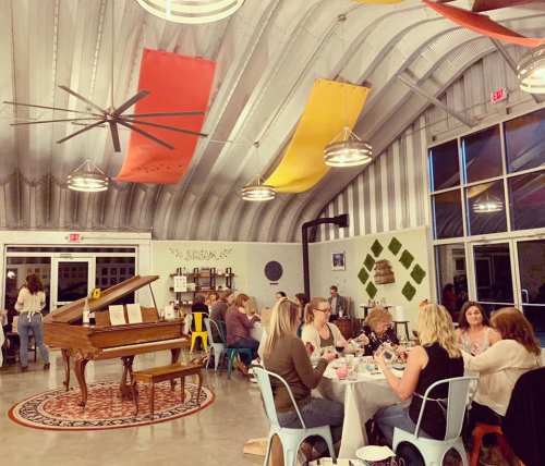 A lively indoor space with a grand piano, colorful banners, and people gathered around tables enjoying an event.