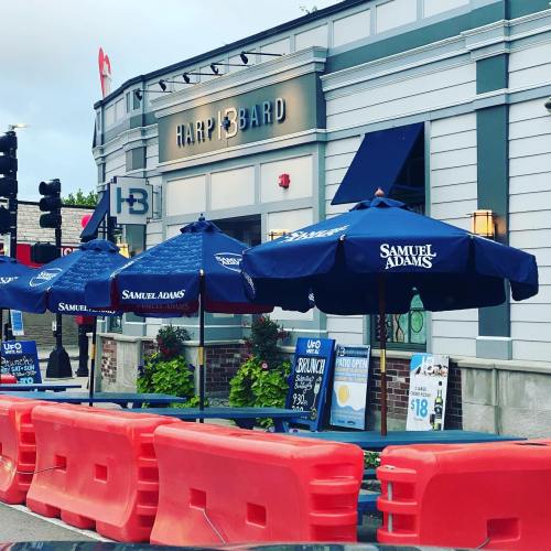 Outdoor seating area of a bar with blue umbrellas, construction barriers, and signage for Samuel Adams and Harp & Bard.