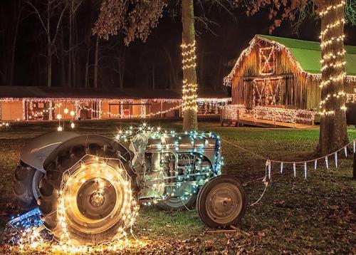 A decorated tractor with colorful lights in front of a barn, surrounded by trees adorned with festive lighting at night.