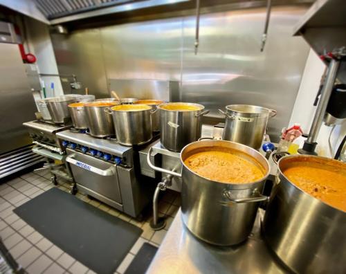 A commercial kitchen with multiple large pots of simmering sauces on a stove, stainless steel surfaces, and cooking equipment.
