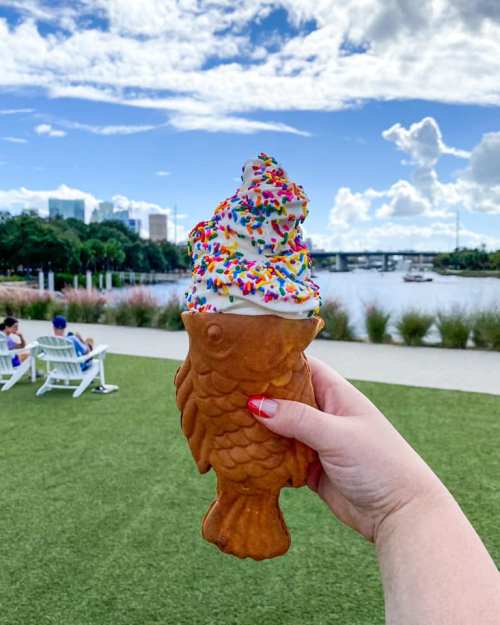 A hand holds a fish-shaped ice cream cone topped with whipped cream and colorful sprinkles, with a scenic background.