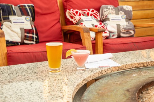 A beer and a pink cocktail sit on a granite table, with cozy blankets and red chairs in the background.