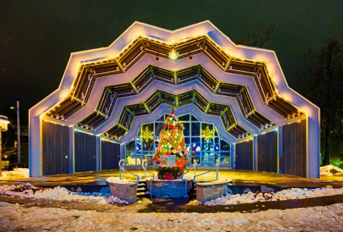 A modern, illuminated building with a unique roof design, featuring a decorated Christmas tree in front, surrounded by snow.