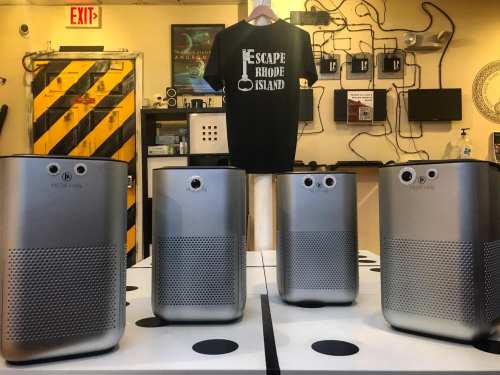 Four silver media devices are lined up on a table, with a black "Escape Rhode Island" t-shirt hanging above them.
