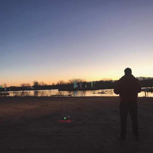 A person stands by a lake at sunset, with a drone on the ground and festive lights reflecting on the water.