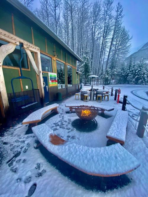 A snowy outdoor seating area with a fire pit, surrounded by trees and a green building in the background.