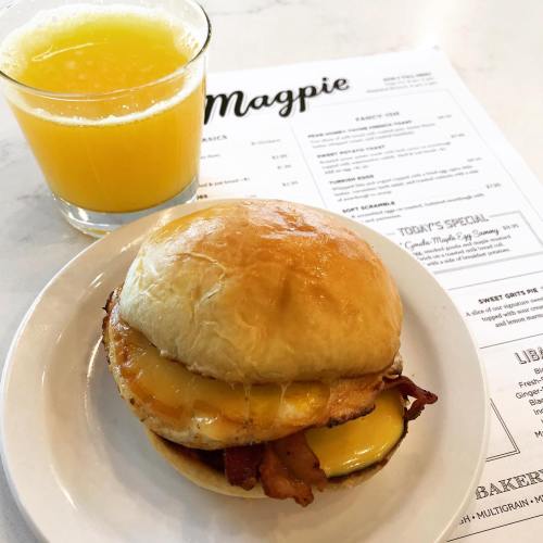 A breakfast sandwich with bacon and cheese on a bun, served with a glass of orange juice and a menu in the background.