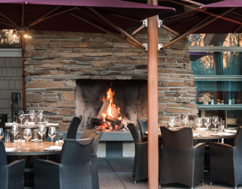 Cozy restaurant setting with a stone fireplace, outdoor tables, and elegant glassware under maroon umbrellas.