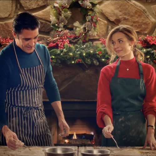 A man and woman in festive attire bake together in a cozy kitchen decorated for the holidays.