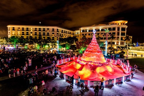A large sand Christmas tree illuminated with colorful lights, surrounded by a festive crowd at night.