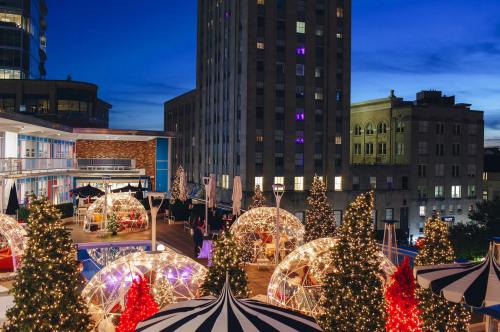 A rooftop scene at dusk featuring illuminated holiday igloos and decorated trees against a city skyline.