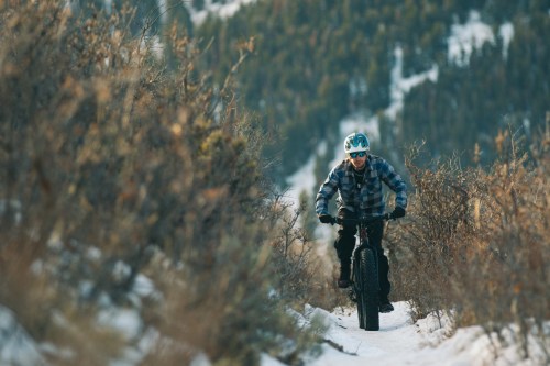 A cyclist rides a fat bike on a snowy trail surrounded by shrubs and trees in a mountainous landscape.