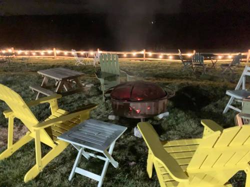 A cozy outdoor setting at night with a fire pit surrounded by colorful chairs and string lights in the background.