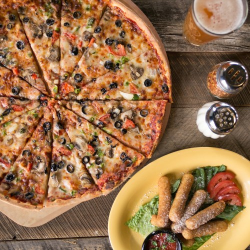 A large pizza topped with vegetables and olives, served with a plate of breadsticks and marinara sauce, alongside a beer.