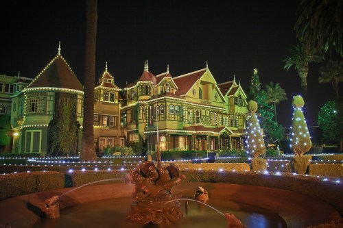 A beautifully lit Victorian mansion at night, surrounded by palm trees and festive decorations.