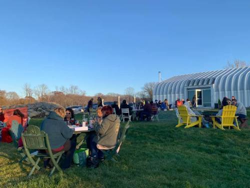 People enjoying a gathering outdoors at tables, with a modern building and clear blue sky in the background.