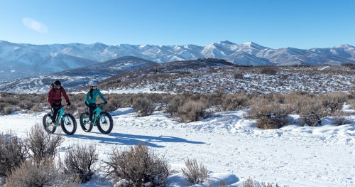 Two cyclists ride fat bikes on a snowy trail with mountains in the background under a clear blue sky.
