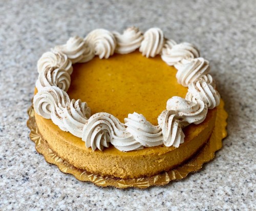 A round pumpkin cheesecake topped with swirls of whipped cream and a sprinkle of cinnamon on a gold cake board.