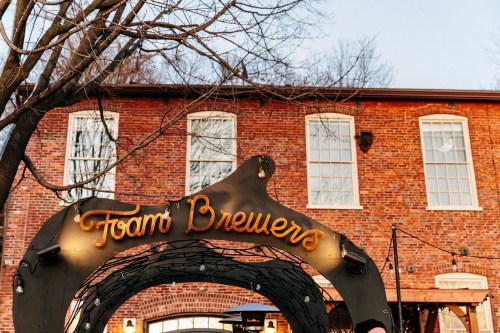 Sign for "Foam Brewers" in front of a brick building, with trees and string lights in the background.