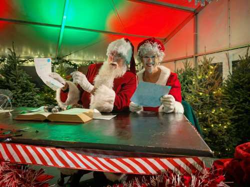 Santa and Mrs. Claus sit at a table, smiling and holding letters, surrounded by Christmas trees and festive decorations.