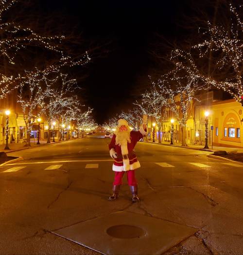 A person dressed as Santa stands in a deserted, festively lit street at night, surrounded by glowing trees.