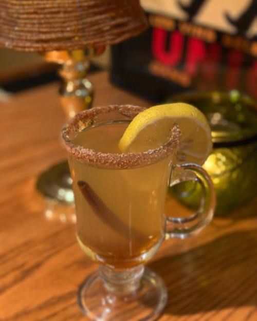 A glass mug filled with a warm drink, garnished with a lemon slice and a cinnamon stick, on a wooden table.