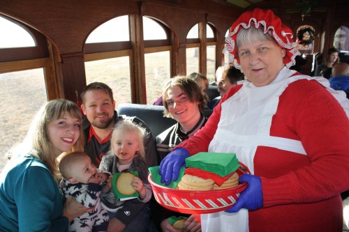 A family on a train with a woman in a holiday costume serving cookies from a tray.