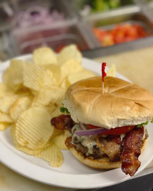 A juicy burger with bacon, lettuce, tomato, and onion, served with a side of crispy potato chips on a white plate.