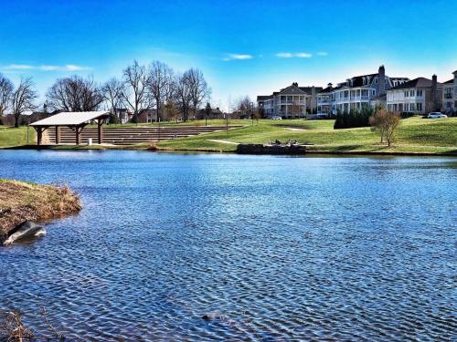 A serene lake view with a pavilion, grassy banks, and elegant homes under a clear blue sky.