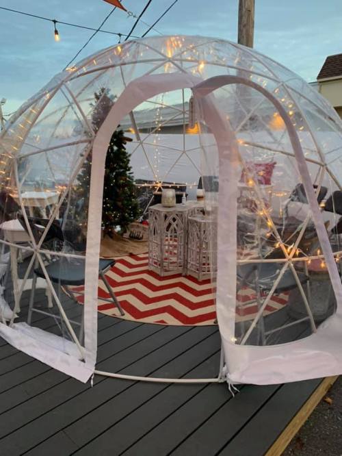 A clear igloo-style structure with string lights, set on a deck with a festive tree and patterned rug inside.