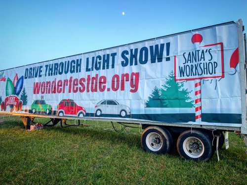 Banner for a drive-through light show featuring "Santa's Workshop" and event details, set against a clear sky.
