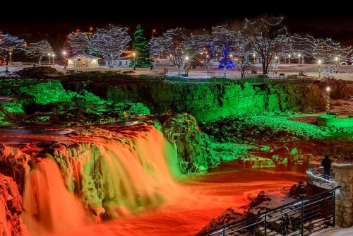 A waterfall illuminated in red and green lights, surrounded by snowy trees and festive decorations at night.