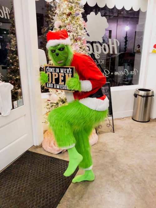 A green Grinch in a Santa outfit holds a sign that says "Come in, we're open and awesome!" near a decorated Christmas tree.