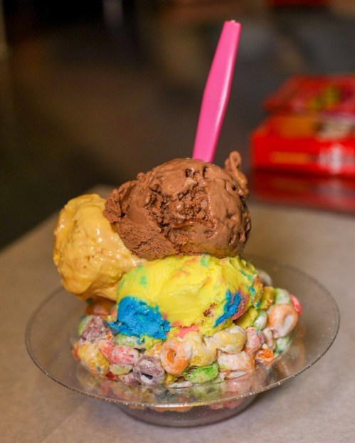 A colorful ice cream sundae with three scoops on a bed of fruity cereal, topped with a pink spoon.