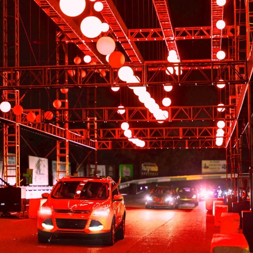 A car drives through a brightly lit, red-themed structure at night, with hanging lights illuminating the scene.