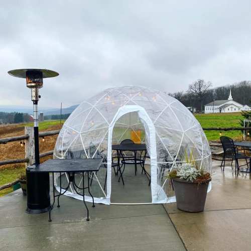 A clear igloo-style tent with tables and chairs, set outdoors on a cloudy day, with a heater nearby.