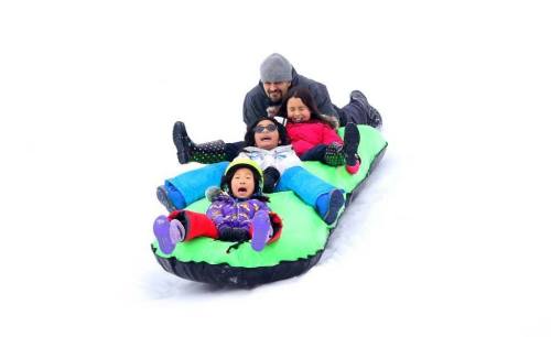 A group of four children and an adult joyfully sledding down a snowy hill on a green sled.