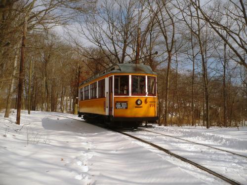 A vintage yellow streetcar on snowy tracks, surrounded by bare trees in a winter landscape.