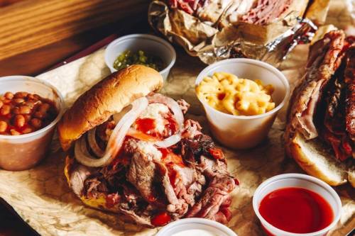 A hearty sandwich filled with sliced meat, topped with onions and sauce, accompanied by sides of macaroni and beans.