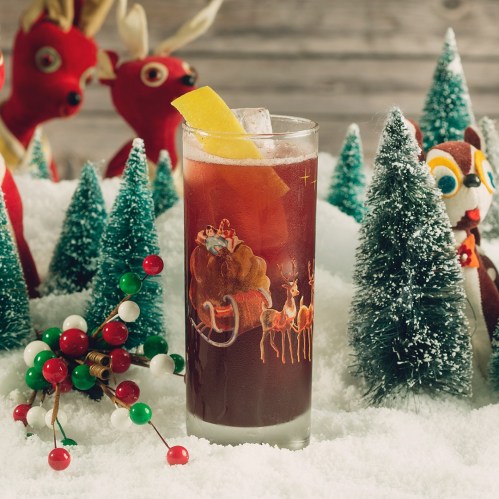 A festive cocktail with a lemon garnish, surrounded by miniature Christmas trees and playful holiday decorations.