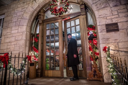 A man in a black coat stands by a beautifully decorated entrance with holiday wreaths and lights.