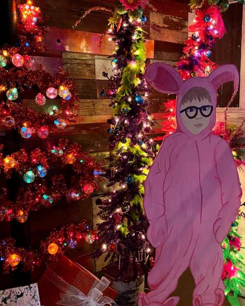 A pink-costumed figure stands beside colorful, decorated Christmas trees with bright lights and ornaments.