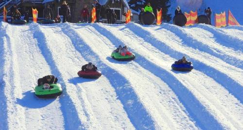 A snowy hill with people sledding down on colorful tubes, surrounded by festive decorations and spectators.
