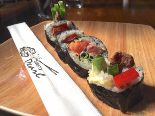 A wooden plate with three sushi rolls, garnished with green onions and a red pepper, beside a branded napkin.