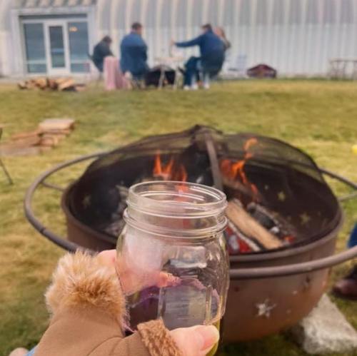A hand holding a jar near a fire pit, with people gathered around in the background.