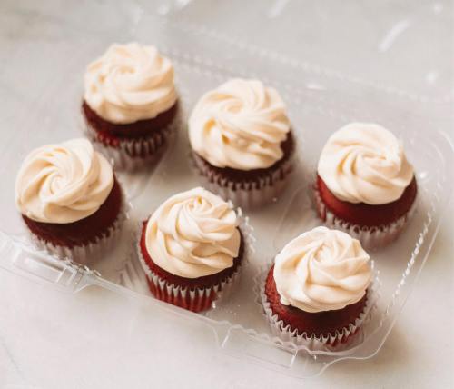 A clear container holding six red velvet cupcakes topped with swirled cream cheese frosting.