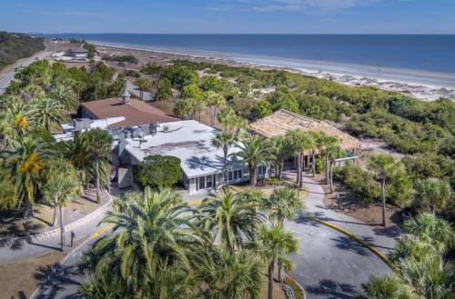 Aerial view of a coastal building surrounded by palm trees, with a sandy beach and ocean in the background.