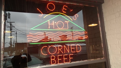 Neon sign in a window reading "Joe's Hot Corned Beef" with colorful graphics of waves and a sun.