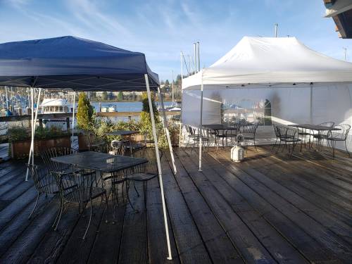 Two tents on a wooden deck overlooking a marina, with empty tables and chairs under clear blue skies.